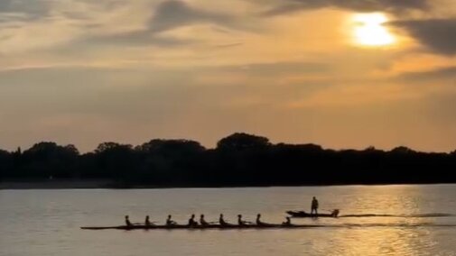 A rowing shell of 8 members at sunrise with teh coach launch following.